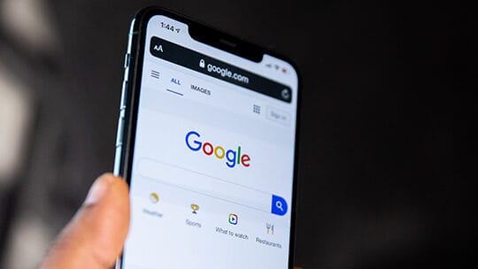 Smartphone displaying the Google search page with a finger holding the side, against a dark background.
