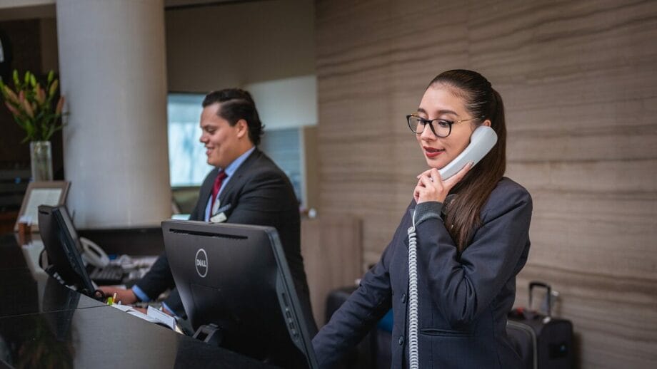A woman in a business suit talking on the phone at a hotel reception desk.