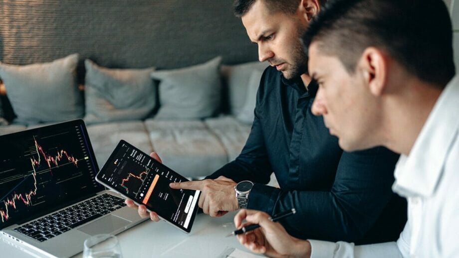 Two men examining financial charts on a tablet and a laptop in a modern workspace setting.