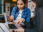 Two people sitting and pointing at a laptop screen, engaged in discussion. One wears a denim jacket and headscarf, the other wears a black top. Bookshelves are visible in the background.