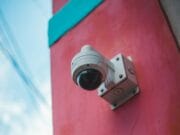 A white security camera is mounted on a red and teal-painted wall, facing slightly downward. The sky with some cables is visible in the background.