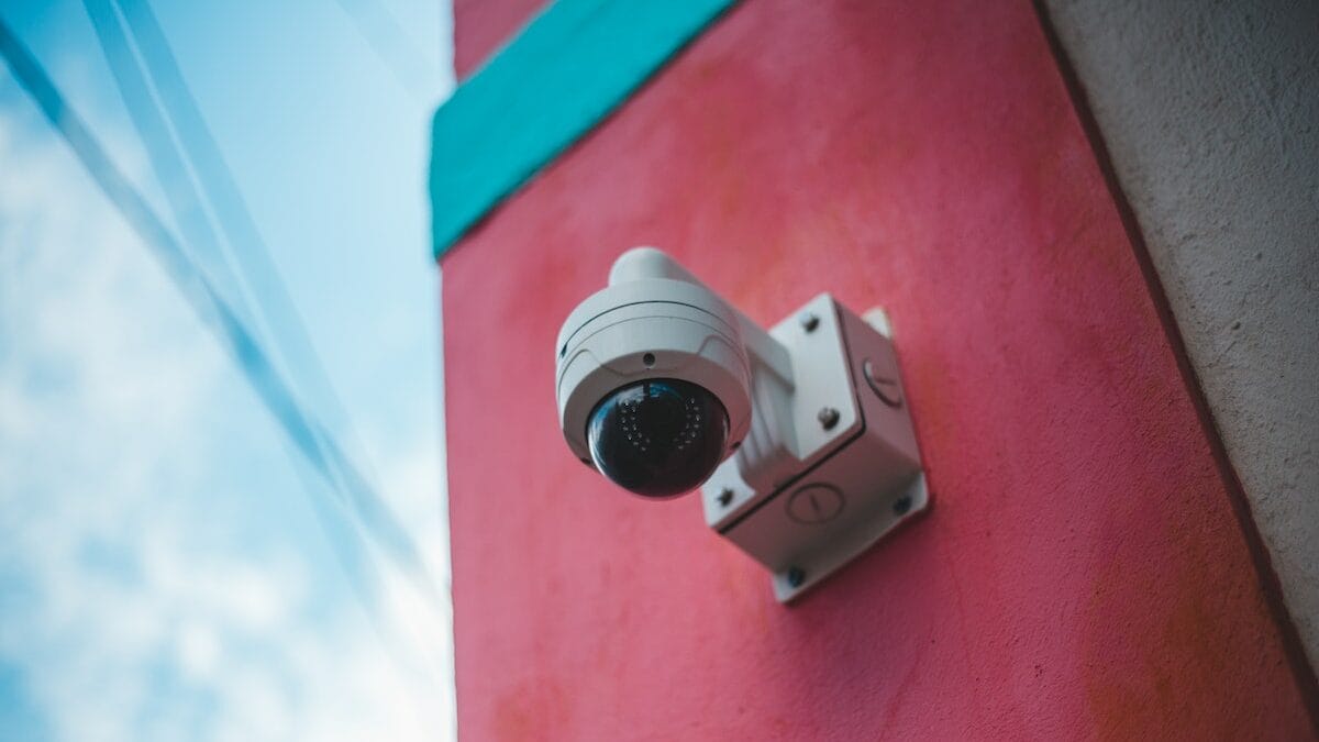 A white security camera is mounted on a red and teal-painted wall, facing slightly downward. The sky with some cables is visible in the background.