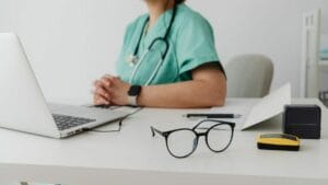 A person in green scrubs and a stethoscope sits at a desk with a laptop, a pair of glasses, a pen, papers, and a small yellow and black device.