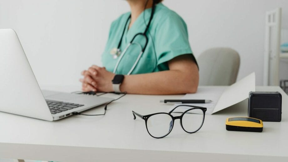 A person in green scrubs and a stethoscope sits at a desk with a laptop, a pair of glasses, a pen, papers, and a small yellow and black device.