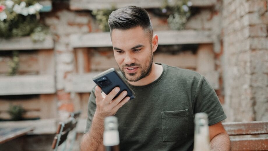 A man is looking at his phone and talking using the speaker while sitting at a table.