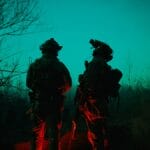 Two soldiers in tactical gear stand outside at night, illuminated by green and red lights, with bare trees in the background.