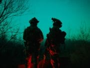 Two soldiers in tactical gear stand outside at night, illuminated by green and red lights, with bare trees in the background.