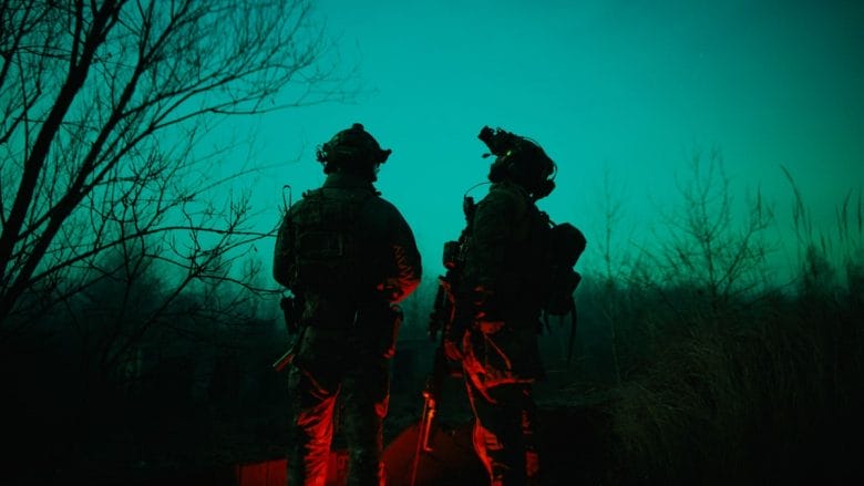 Two soldiers in tactical gear stand outside at night, illuminated by green and red lights, with bare trees in the background.