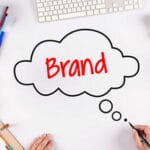 A person draws a thought bubble with the word Brand on a white desk surrounded by office and art supplies.