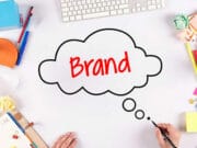 A person draws a thought bubble with the word Brand on a white desk surrounded by office and art supplies.
