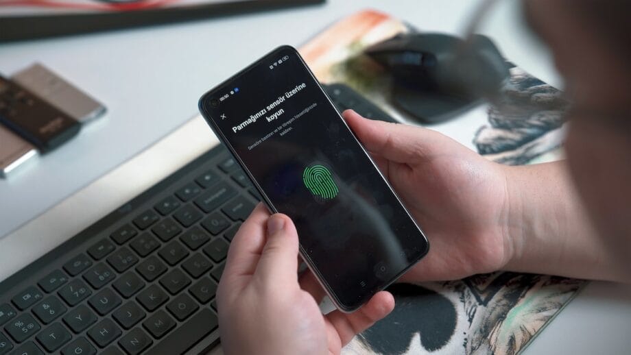 Person holding a smartphone with a fingerprint sensor setup screen displayed. A keyboard and other items are visible in the background.