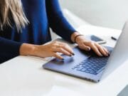 A woman in blue long sleeve shirt typing on a laptop at a desk.