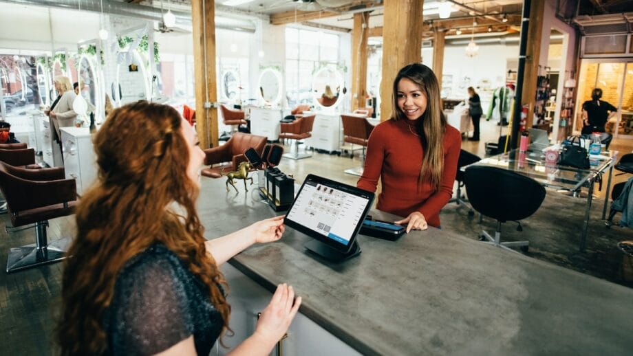 Two woman at a hair salon with a tablet in hand interacting with each other.