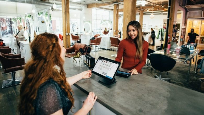 Two woman at a hair salon with a tablet in hand interacting with each other.