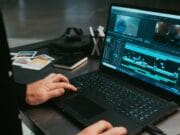 Person using a laptop with video editing software open, surrounded by notebooks, photos, and electronic accessories on a desk.