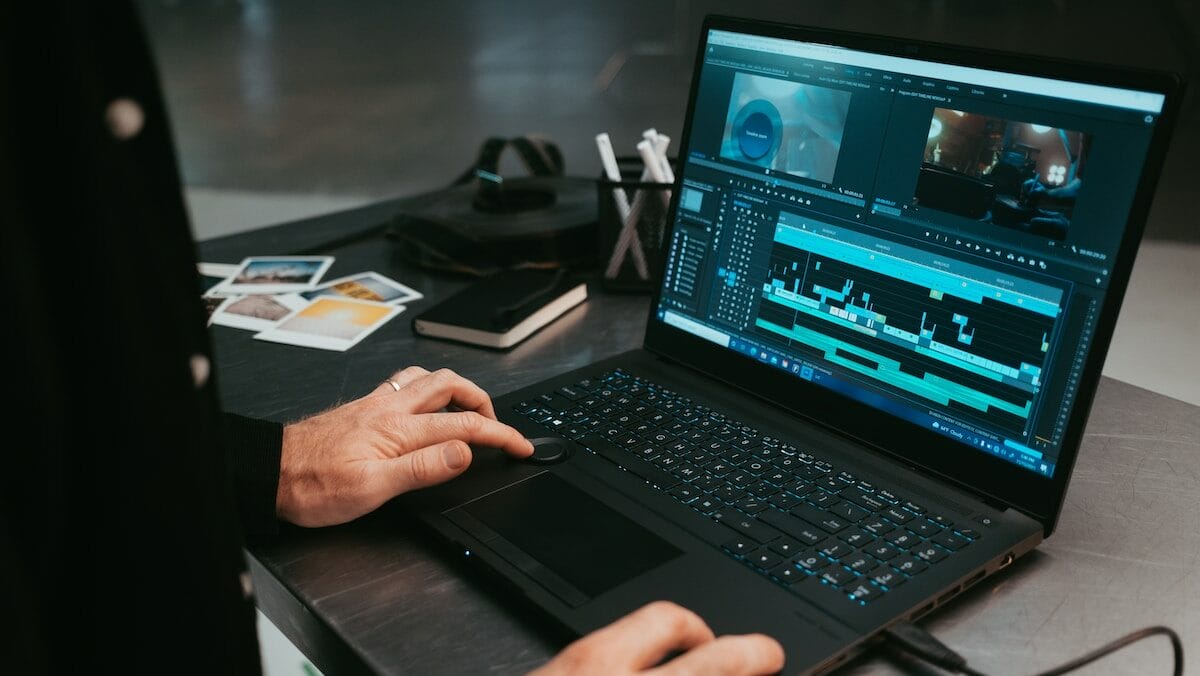 Person using a laptop with video editing software open, surrounded by notebooks, photos, and electronic accessories on a desk.