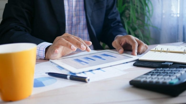 Person in a suit reviews bar chart on a tablet at a desk; nearby are a yellow mug, planner, pen, and calculator.