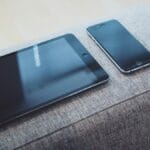 A gray couch with a tablet and phone on it.