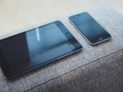 A gray couch with a tablet and phone on it.