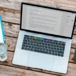 A laptop on a wooden table with a notebook and phone.