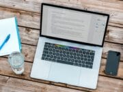 A laptop on a wooden table with a notebook and phone.