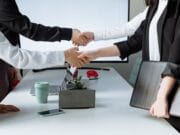 Four individuals shaking hands over a table in an office environment, suggesting a professional agreement or greeting.