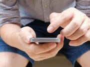 A person seated on a wooden bench is using a smartphone with both hands.