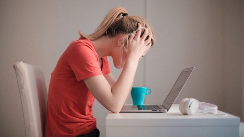 A person sitting at a desk with their head in their hands, appearing stressed or upset, in front of an open laptop with a blue cup and white headphones nearby.