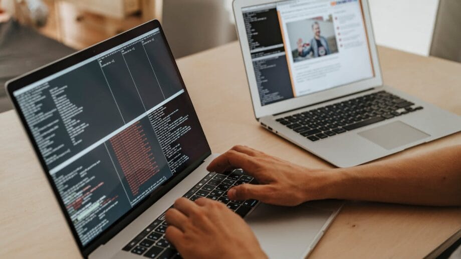 A person types on a laptop with code on the screen while another laptop displays a web page.