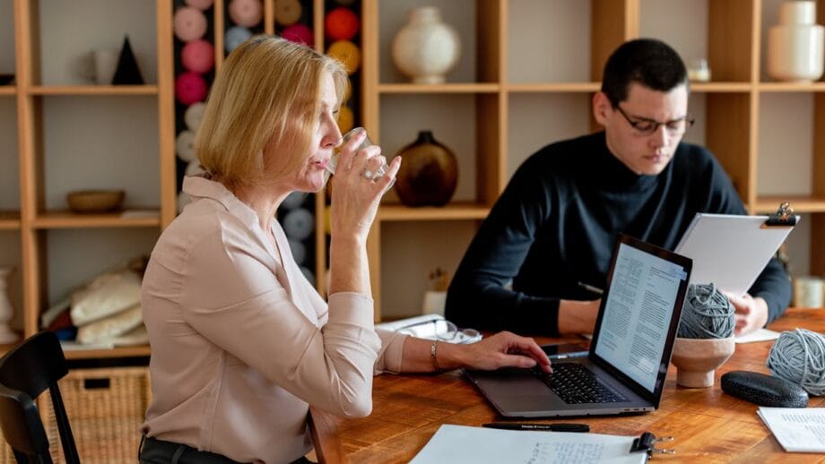 A woman sipping a drink while sitting at a table with a laptop, with a man next to her focused on paperwork in a room with shelves of yarn and pottery.