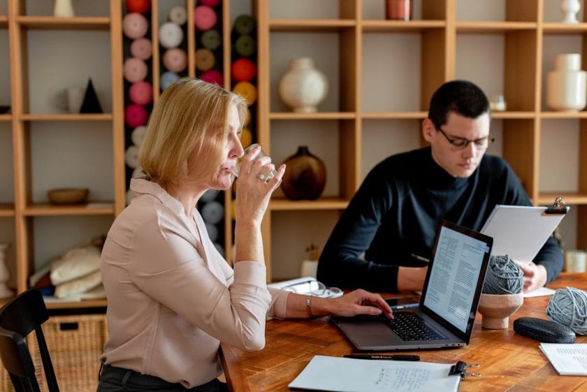 A woman sipping a drink while sitting at a table with a laptop, with a man next to her focused on paperwork in a room with shelves of yarn and pottery.