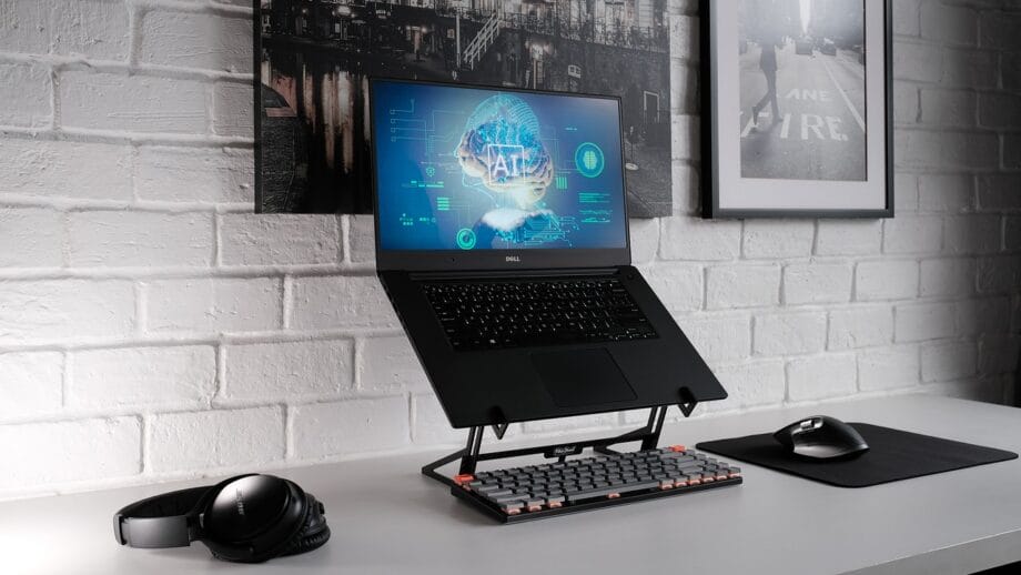 A modern, minimalist workspace featuring a laptop on a stand, a mechanical keyboard, a mouse on a pad, and headphones, against a white brick wall backdrop with framed art.