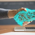 A person's hand extends towards a laptop screen with a graphic of a blue, geometric handshake appearing, symbolizing digital connectivity or online agreements.