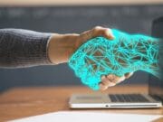 A person's hand extends towards a laptop screen with a graphic of a blue, geometric handshake appearing, symbolizing digital connectivity or online agreements.