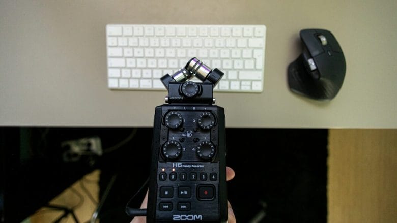 A hand holding a zoom h5 audio recorder with a crossed pair of microphones above a white keyboard and a black computer mouse on a desk.