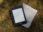 An e-reader device with a visible screen displaying text, resting on top of a gray felt cover, lies on a natural surface covered with grass and moss.