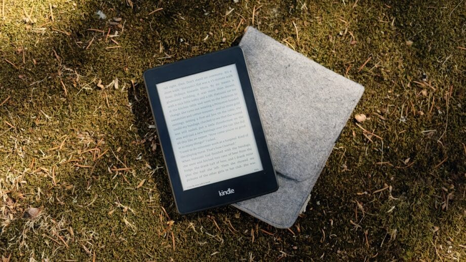 An e-reader device with a visible screen displaying text, resting on top of a gray felt cover, lies on a natural surface covered with grass and moss.