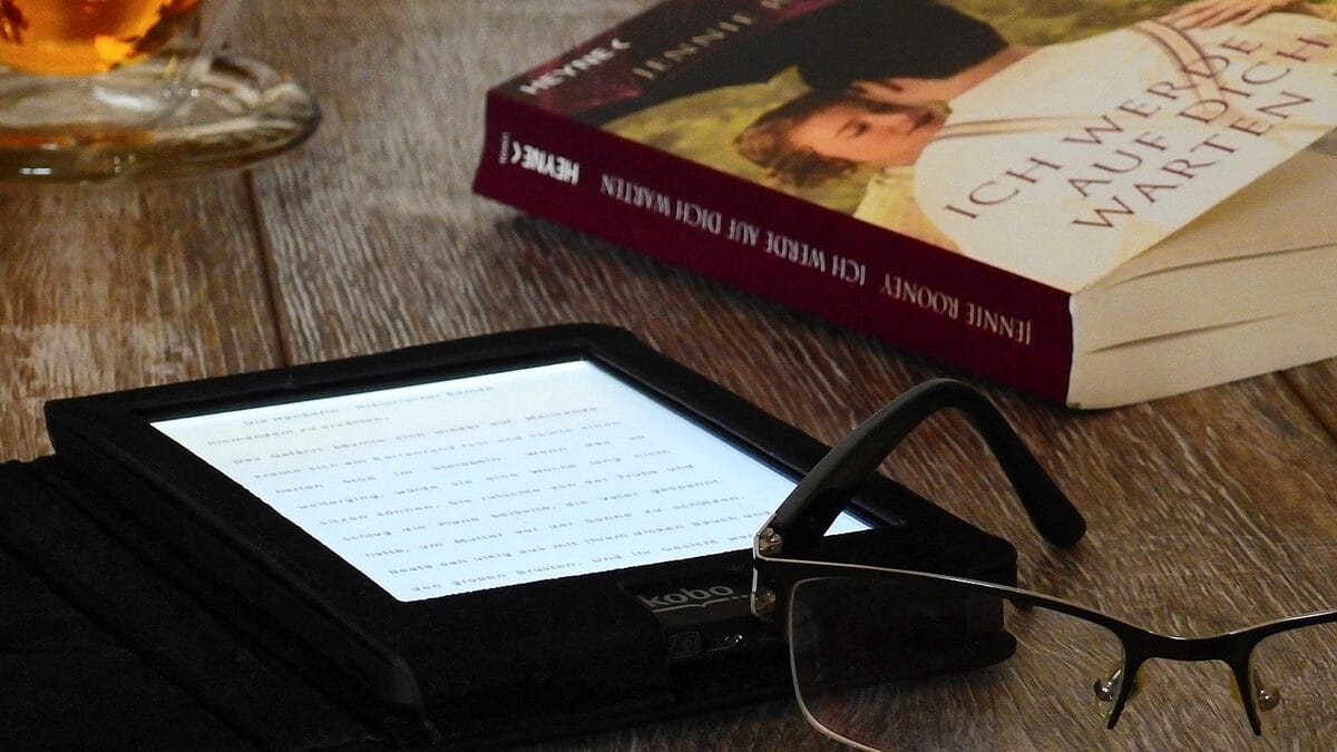 A hardcover book, e-reader, eyeglasses, and a cup of tea arranged on a wooden surface.