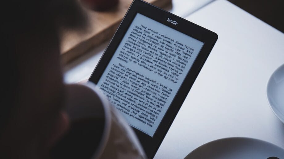 An e-reader displaying text rests on a table beside a cup of coffee and a partially visible plate, suggesting a moment of leisure or study.