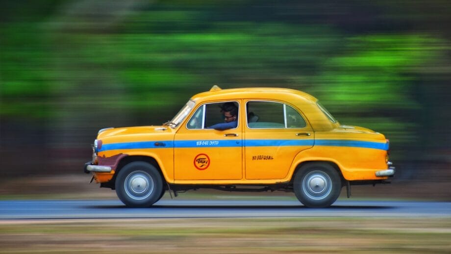 A yellow taxi with black stripes and the word "taxi" printed on it is in motion on a blurred street, indicating a panning photography technique to capture the sense of speed.