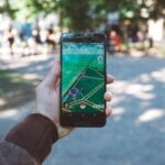 A person's hand holding a smartphone displaying a location-based augmented reality game, with blurred figures and trees in the background in a park setting.