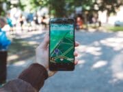 A person's hand holding a smartphone displaying a location-based augmented reality game, with blurred figures and trees in the background in a park setting.