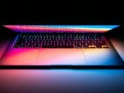 A laptop with a colorful backlit keyboard open in a dimly lit room, emitting a soft glow onto the surrounding surface.