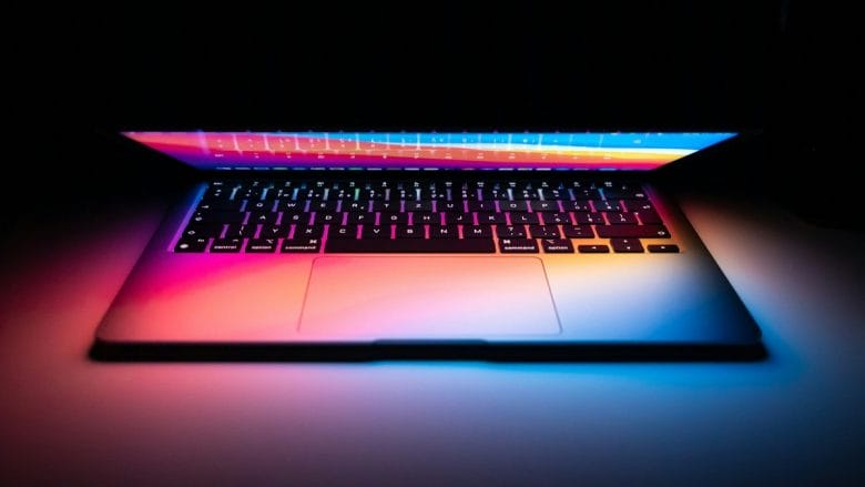 A laptop with a colorful backlit keyboard open in a dimly lit room, emitting a soft glow onto the surrounding surface.
