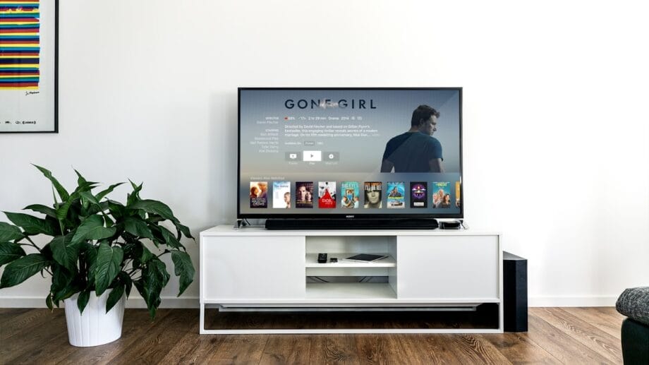 A modern living room features a large flat-screen TV on a white entertainment unit. The TV display shows the movie "Gone Girl" on a streaming service. There's a green potted plant on the left and a colorful abstract painting on the wall.
