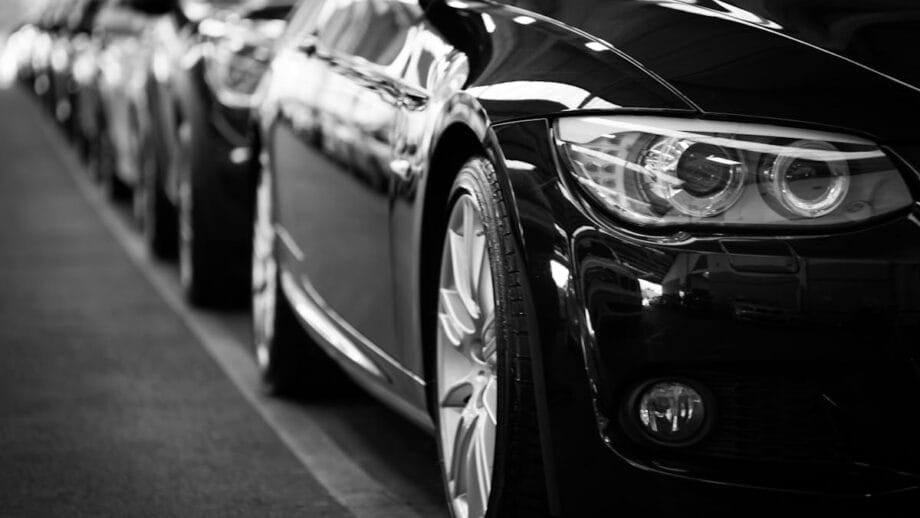 A black and white photo showcasing a row of cars parked in a line, focusing on the shiny, detailed headlights and mirrors of the foremost car.
