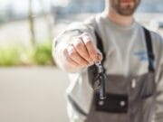 A man in a gray uniform holds out a set of car keys, focusing the camera on the keys with a softly blurred background.