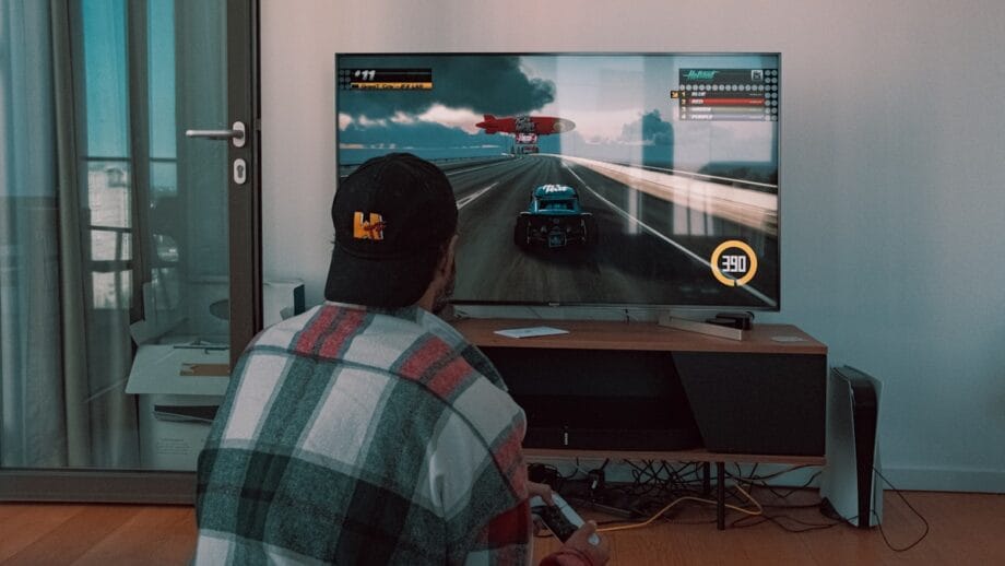 Person wearing a plaid shirt and cap is sitting on the floor playing a racing video game on a television in a living room.