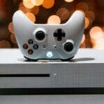 A white Xbox One console with a matching controller, placed on a gray surface. The console has an illuminated power button, and blurred lights are in the background.