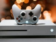 A white Xbox One console with a matching controller, placed on a gray surface. The console has an illuminated power button, and blurred lights are in the background.